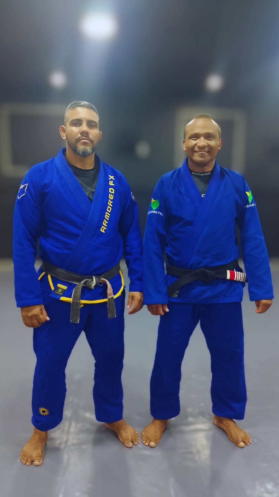 Two athletes in blue ARMORED FX gis with black belts on the mat.